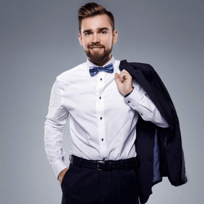 Dapper man wearing a white shirt and blue bow tie, holding a jacket, set against a grey background.